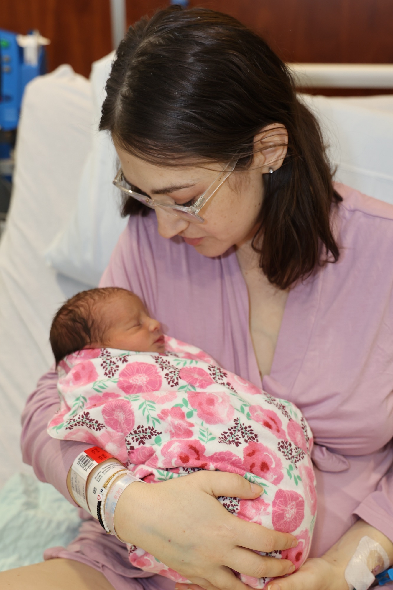 Author sits in hospital bed, wearing a purple robe, holding newborn baby who is wrapped in a pink floral blanket 